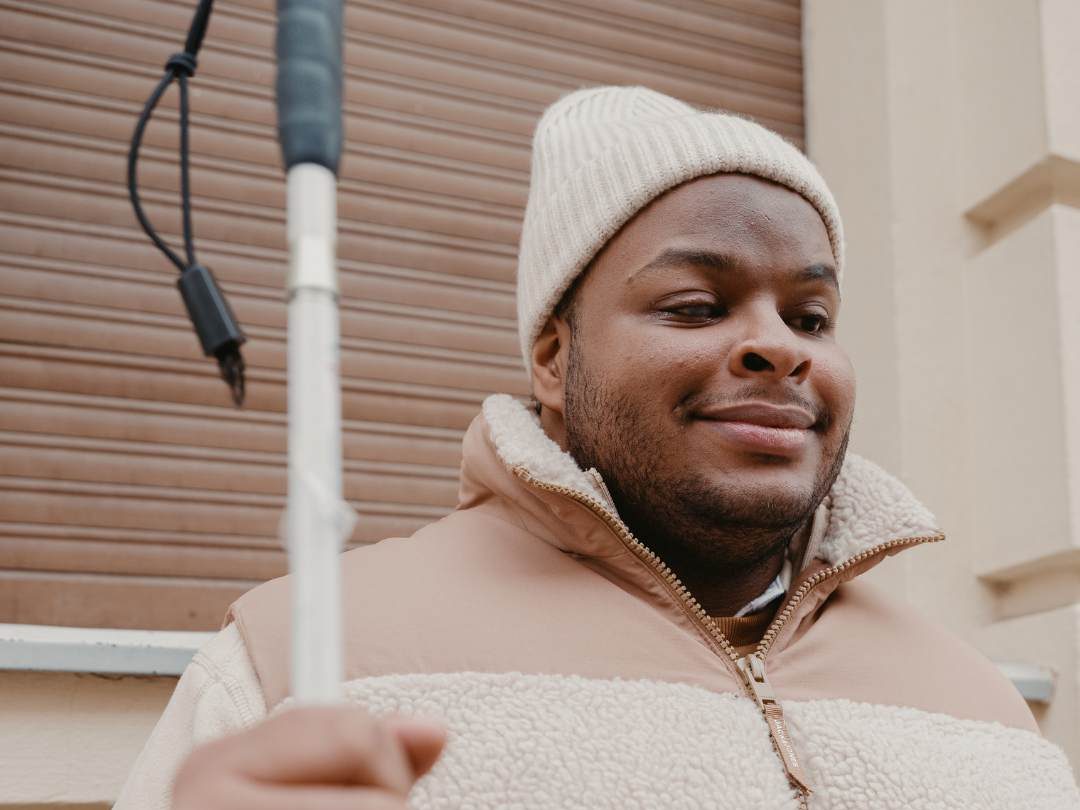 Photo of visually impaired man in a beige fleece vest, and coordinating hat and sweater, holding his cane, and smiling. He is standing in front of a building. 