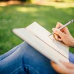  woman outside seated in the grass, taking notes with a notebook pen